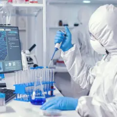 Osoba w kombinezonie ochronnym pracuje z pipetą w laboratorium medycznym, obok monitora z obrazem molekularnym.