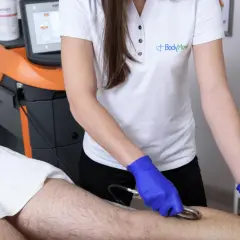 Fizjoterapeuta w niebieskich rękawiczkach wykonuje zabieg na nodze pacjenta za pomocą urządzenia z elektrodą w kształcie gęsiej stopki.