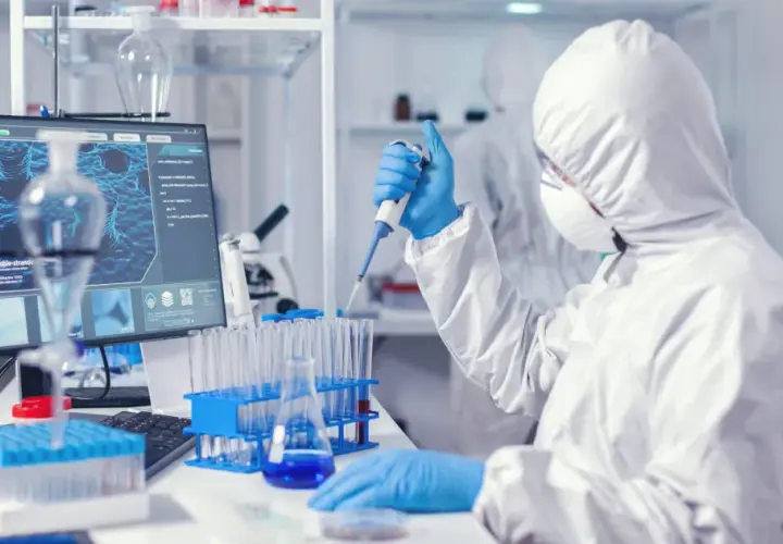 Osoba w kombinezonie ochronnym pracuje z pipetą w laboratorium medycznym, obok monitora z obrazem molekularnym.