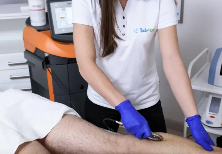Fizjoterapeuta w niebieskich rękawiczkach wykonuje zabieg na nodze pacjenta za pomocą urządzenia z elektrodą w kształcie gęsiej stopki.
