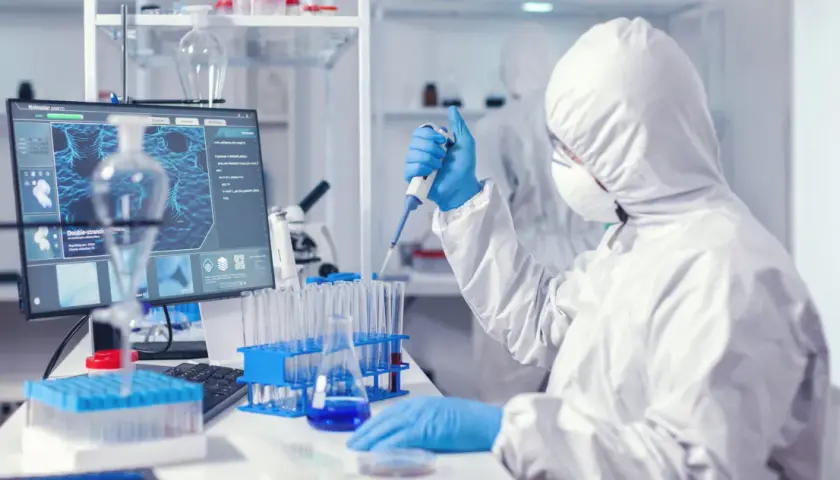 Osoba w kombinezonie ochronnym pracuje z pipetą w laboratorium medycznym, obok monitora z obrazem molekularnym.