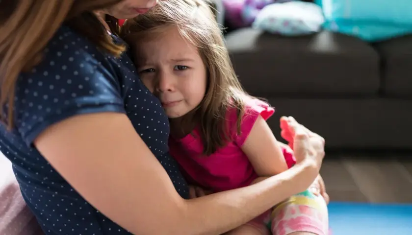 Mała dziewczynka z zapłakaną buzią mocno przytula się do mamy, wyrażając swój lęk separacyjny.