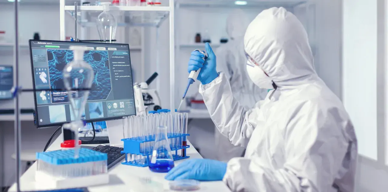 Osoba w kombinezonie ochronnym pracuje z pipetą w laboratorium medycznym, obok monitora z obrazem molekularnym.